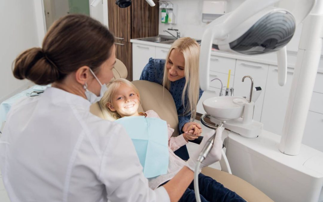 child dental patient in dentist chair, holding mother's hand, smiling at dentist